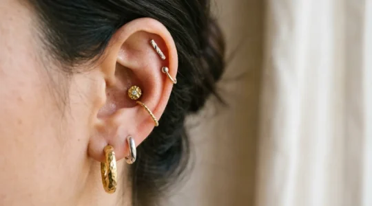 Gros plan artistique sur une oreille avec une composition harmonieuse de bijoux dorés et argentés, montrant un équilibre entre les piercings du lobe, hélix, tragus et conch